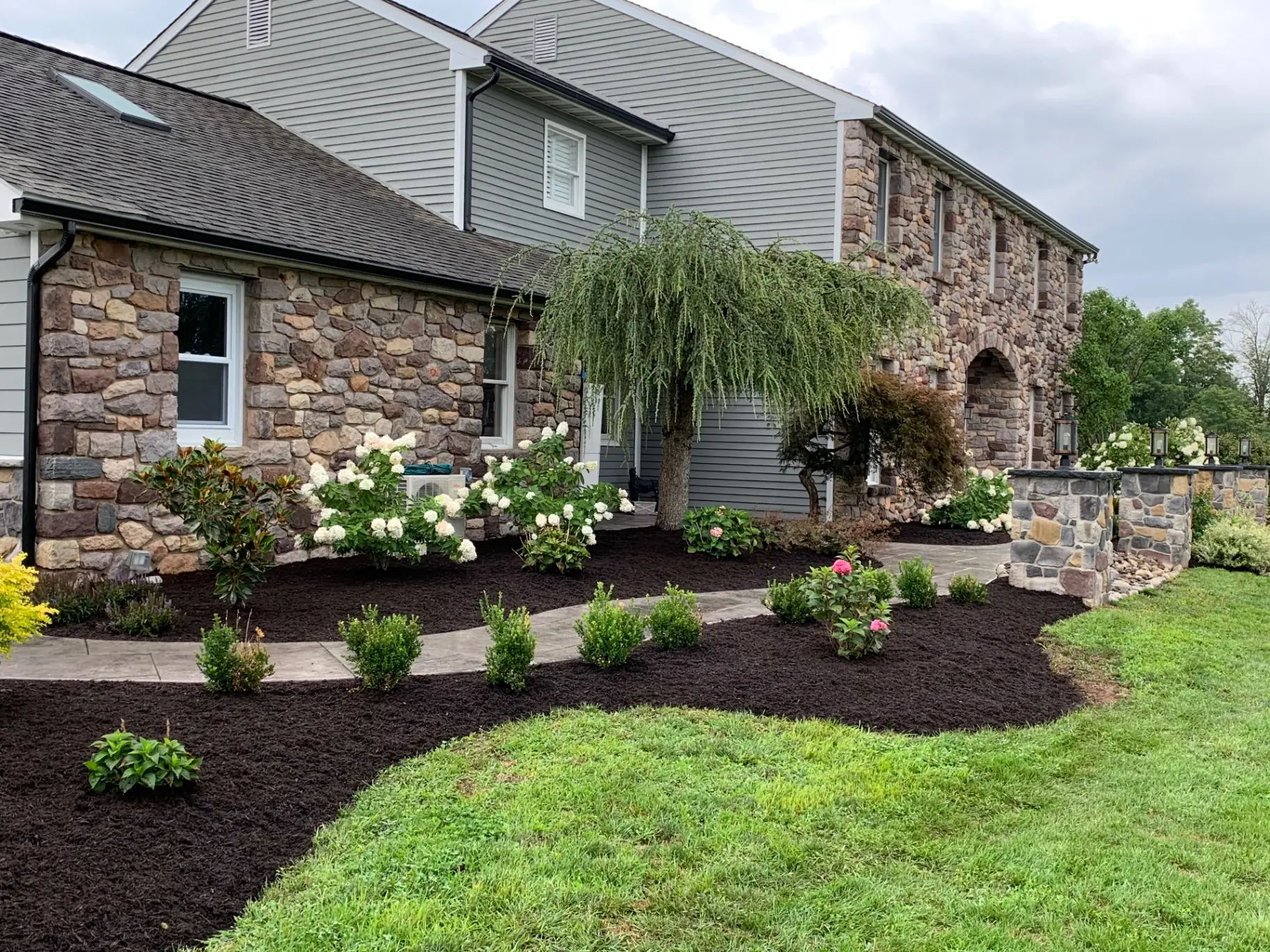 New planting beds with shrubs and flowering plants along a curved stamped concrete walkway beside a stone home in Buckingham, PA